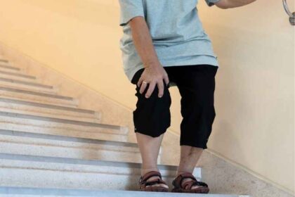 Persona mayor bajando escaleras mientras se sujeta la rodilla con dolor, posible crepitación o desgaste articular.