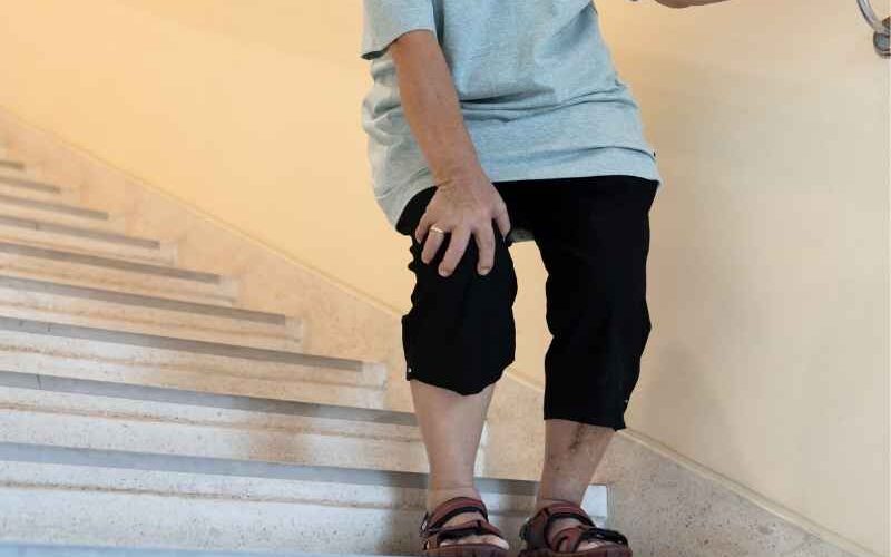 Persona mayor bajando escaleras mientras se sujeta la rodilla con dolor, posible crepitación o desgaste articular.